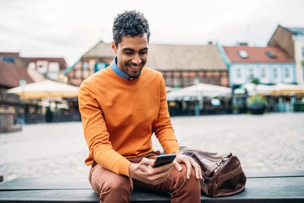 Man Outdoors By The Water Sitting On The Bench With Phone In Hand