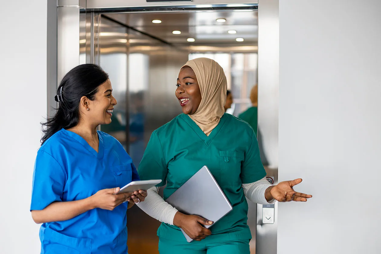 Two female nurses smiling and talking while walking by an elevator.
